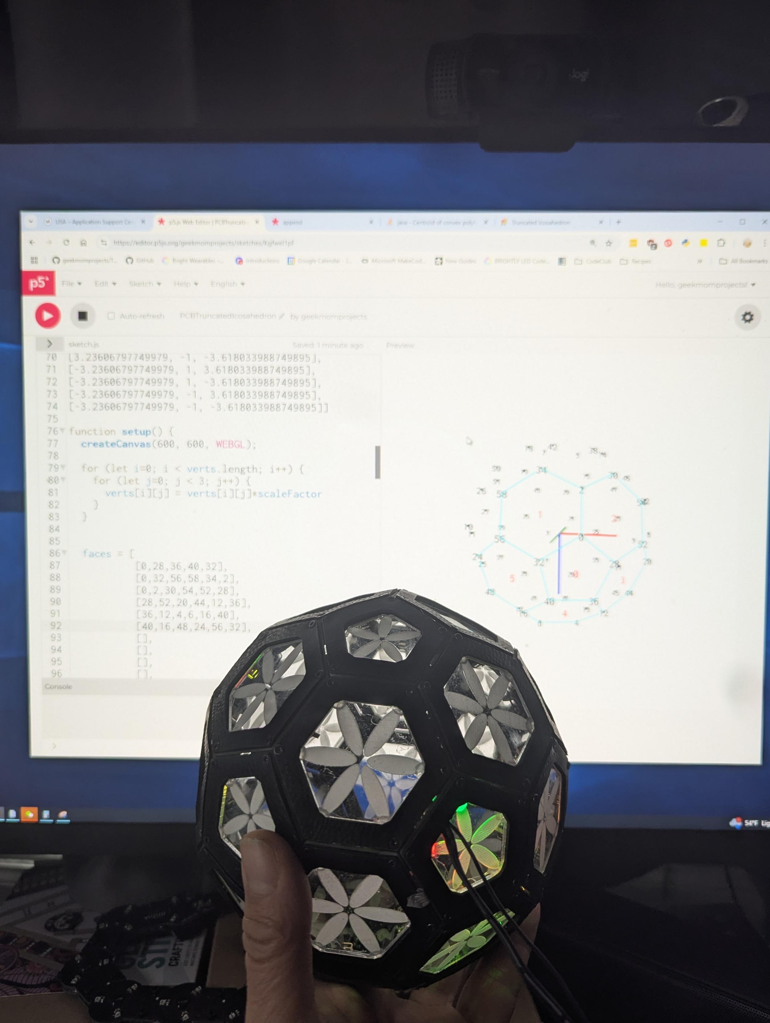 A hand holding a black truncated icosahedron with translucent acrylic flowers inset each side, in front of a computer screen. The screen shows a 3D rendering of labelled points corresponding to each of the vertices of the truncated icosahedron, and a partially completed list of faces comprised of lists of vertices within the code.