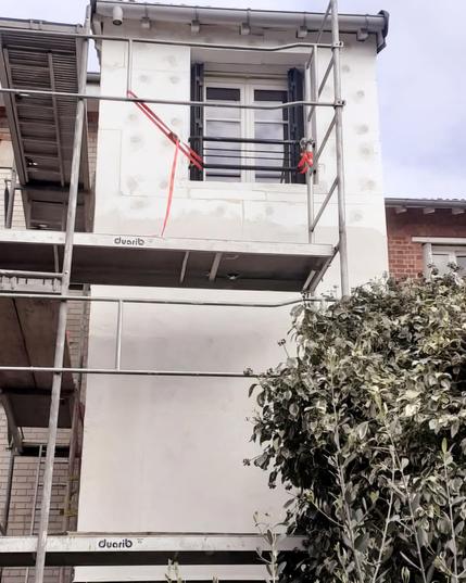 Une façade en cours d'isolation par l'extérieur, l'enduit est en train d'être mis. Il y a une fenêtre en haut, un échafaudage tout du long et un arbuste en bas à droite.