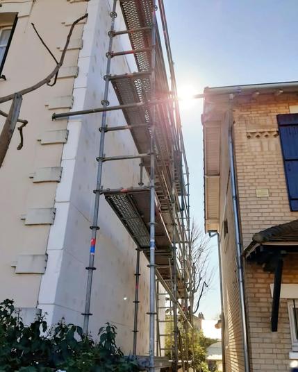Une façade en cours d'isolation par l'extérieur vue de profil, l'enduit est en train d'être mis. Il y a un échafaudage tout du long, un rayon de soleil de soleil en haut, à droite un autre bâtiment qui fait face à la façade et en bas à gauche un arbuste.