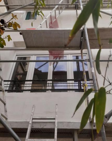 Une façade en cours d'isolation par l'extérieur avec une fenêtre large au milieu, l'enduit est en train d'être mis. Il y a un un échafaudage tout du long des échelles partout et quelques feuilles qui passent devant l'objectif.