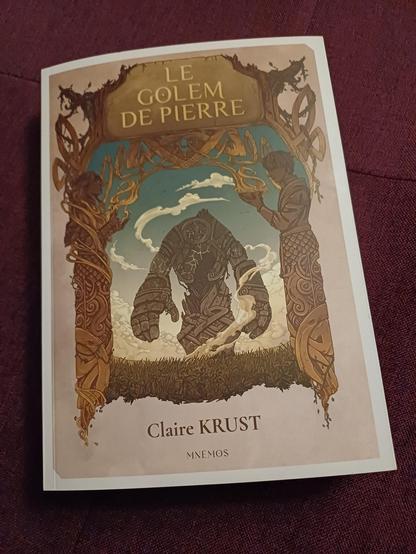 Gros plan de la couverture du roman "Le golem de pierre" de Claire Krust, publié chez Mnemos. On y voit un golem en train de marcher dans une prairie. Il s'approche d'une arche dont les deux piliers sont une femme et un homme. Couverture signée Cyrielle Foucher.