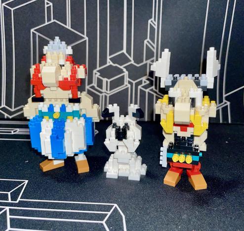 The image features three LEGO-style figurines: a large figure resembling a Viking with a red tunic, a smaller white creature, and another Viking-like figure dressed in yellow and black. They are set against a geometric black background.