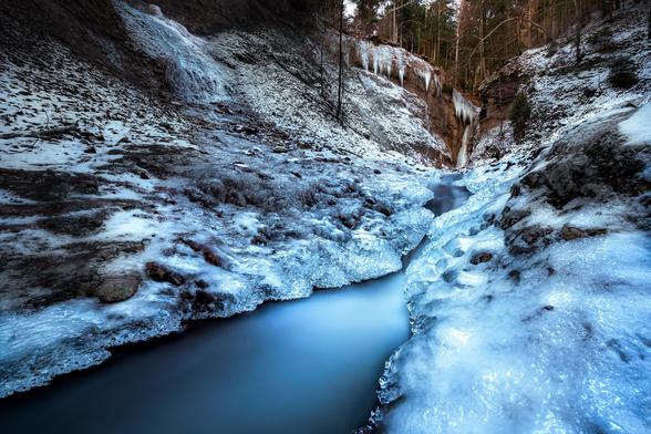 Langzeitfotografie eines von Eis gesäumten Baches. Im Hintergrund ist ein kleiner Wasserfall zu sehen.