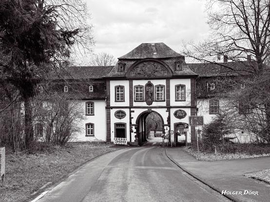 Schwarz-Weiß-Aufnahme des historischen Torhauses des ehemaligen Klosters Arnsburg bei Lich. Das barocke Gebäude mit Rundbogendurchgang, verzierten Fenstern und einer Reliefplatte über dem Tor steht inmitten einer winterlichen Landschaft mit kahlen Bäumen. Die monochrome Farbgebung verstärkt die zeitlose, melancholische Atmosphäre und hebt die architektonischen Details eindrucksvoll hervor.
