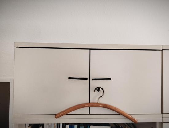 Die obere Hälfte eines Büroschranks. Zwei Türgriffe, an einem hängt ein Kleiderbügel, so dass sich der Eindruck eines traurigen gesichts ergibt