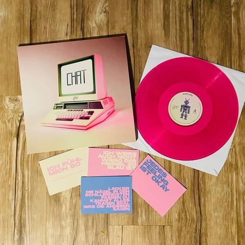 Ein künstlerisches Display mit einer rosa Schallplatte mit dem Titel "CHAT" neben einem farbenfrohen Albumcover, das einen Retro-Computer darstellt. Um das Vinyl herum befinden sich mehrere gedruckte Karten mit deutschem Text, die alle auf einer Holzoberfläche angeordnet sind.