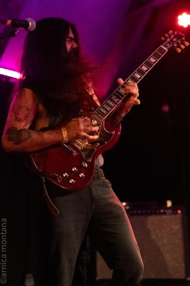 A musician passionately playing a red electric guitar on stage, with long flowing hair and visible tattoos on their arm. The scene is illuminated by colorful stage lights, creating a dynamic atmosphere.