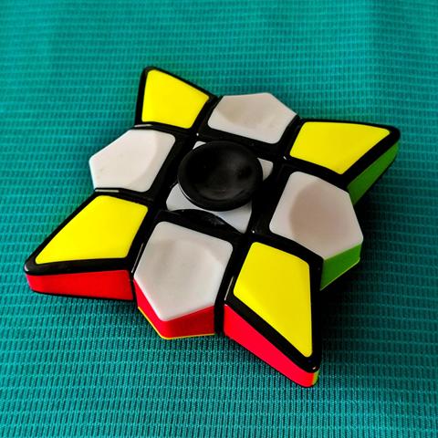 Foto del cubo spinner en versión spinner.
Es como un cubo de Rubik al que le has quitado la capa superior e inferior. En lugar de tener lados lisos, tienen formas más puntiagudas.
En el centro de las caras blanca y amarilla tiene un círculo negro (habría estado bien que fuese del color de la cara) que actúa como base sobre la que gira.