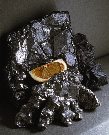 A still life composition of dark, metallic, rock-like formations with a dried orange slice resting among them. The textures and contrast evoke an abstract vision of the planet Mercury—harsh, reflective, and touched by fleeting warmth.