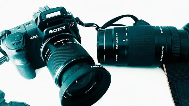 The image shows a Sony α200 DSLR camera with a lens attached, along with an additional Sony zoom lens placed next to it. The camera is black with a textured grip, and the brand name "Sony" is clearly visible on the front. The lens mounted on the camera has a focal length range of 18-70mm, while the separate telephoto zoom lens has a focal length range of 75-300mm. This setup is a photography kit suitable for different types of shots, from wide-angle to long-distance zoom. The background is light-colored, and the image has a cool-toned filter applied, giving it a bluish hue.