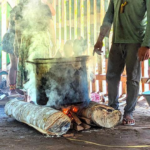 A large pot boiling over a fire, with a Huni Kuin man nearby watching over it.