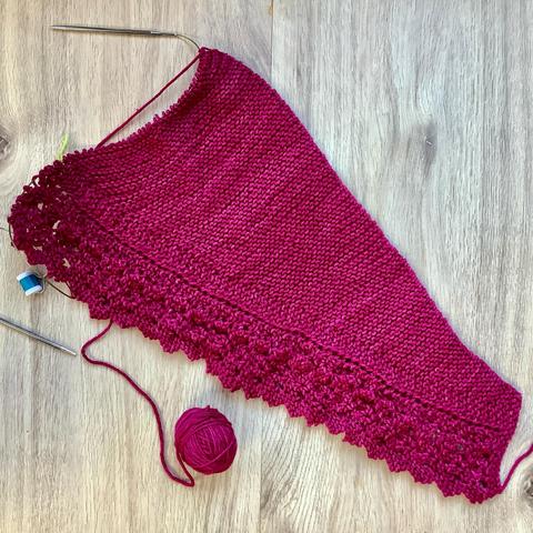 Flat lay view of a knitting project, featuring garter stitch and lace on a circular needle. It's shown on a light wooden background.