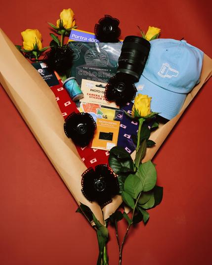 A creatively arranged photography-themed gift bouquet wrapped in brown paper, featuring yellow roses, a blue '400 ISO' embroidered cap, a Tamron camera lens, a roll of Portra 400 film, a photographer's coloring book, and various photography-related accessories. The bouquet also includes socks with camera patterns, sticker packs, an enamel pin, and decorative black film reel flowers. The background is a warm red tone, complementing the aesthetic presentation.
