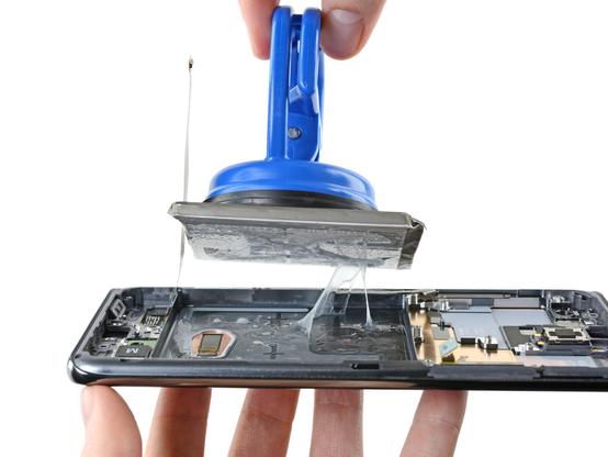 Galaxy S20 Ultra with its back cover and most of its internals removed. The battery is being held directly above its cut-out in the frame of the phone by a heavy duty suction cup. The battery is currently being removed, evidenced by the thick glue on the battery and phone itself, strands of which are attached between them.