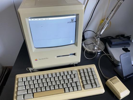 Picture of an Apple Macintosh Plus computer with keyboard and mouse on a black desk. The computer is turned on, and the desktop system can be seen on the screen.
