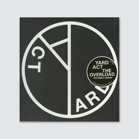 This is the album cover for The Overload, the debut album by Yard Act. The design features a bold, black-and-white minimalist aesthetic with a large, circular emblem that appears to be a stylized version of the band's name, split into geometric sections. The typography is simple and modern, with "YARD ACT" and "THE OVERLOAD" highlighted in a sticker-like label on the right side. The stark contrast and structured design give the cover a striking, almost post-punk or industrial feel, reflecting the band's sharp, satirical style.