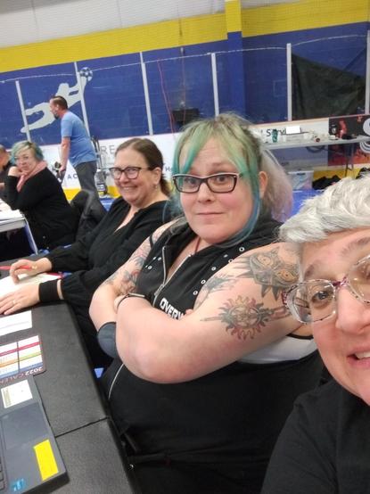 Several humans of various genders smiling at the camera. They are sitting at a table keeping score for a roller derby game.