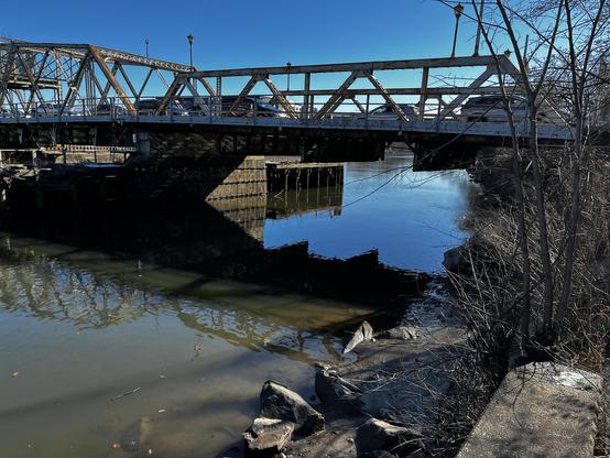 The Lower Passaic river, its water urban brown, lies smooth beneath the De Jessa bridge in Nutley, New Jersey. A car wheel is faintly visible in the sediment in the lower left quadrant. Exposed tidal mud hosts an array of largish stones on the right. The complexity of the bridge tries to dominate the upper portions of the photograph, but the sky and the river are too much for it.