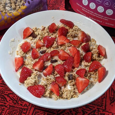 A white bowl filled with granola, raisins, and fresh sliced strawberries, partially soaked in milk or a smoothie base. The bowl sits on a red-patterned surface, with a bag of granola and a protein supplement package visible in the background. The vibrant red strawberries contrast with the golden-brown granola, creating a visually appealing and nutritious meal.
