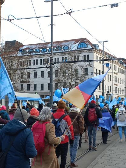 Ein Demonstrationszug läuft von der Kamera weg, sodass die Teilnehmenden alle von hinten zu sehen sind. Viele tragen hellblaue Fahnen oder Ballons, auf denen eine weiße Taube abgebildet ist. Eine grauhaarige Demonstrantin mit rotem Rucksack trägt eine Fahne, die aus der deutschen und der russischen Flagge zusammengesetzt ist. Über den Demonstrierenden verlaufen Oberleitungen, im Hintergrund ist ein mehrstöckiges Gebäude zu sehen.