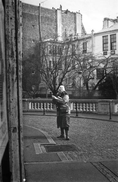 Photo en noir et blanc d’une femme debout devant l’Église Saint-Vincent-de-Paul, ajustant son appareil photo. Elle porte une veste chaude et une jupe longue, concentrée sur son cadrage. Autour d’elle, le décor urbain se déploie : façades anciennes, arbres dénudés et pavés sous ses pieds. Comme dans ses dessins sur papier noir et encre blanche, elle joue avec les contrastes, sculptant la lumière à travers son objectif.