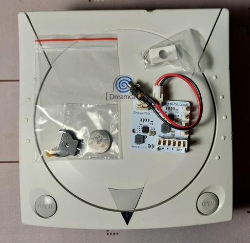 Man sieht eine Draufsicht auf eine Sega Dreamcast. Auf der Dreamcast liegt in einer kleinen Plastiktüte ein Knopfzellen-Akku, eine Batteriehalterung und eine Sicherung. Rechts daneben liegt ein DreamPSU Netzteil, das das Originalnetzteil der Sega Dreamcast ersetzen soll.