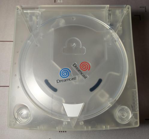 Man sieht die Draufsicht eines transparenten Sega Dreamcast Gehäuses. Auf dem Gehäuse liegen zwei Dreamcast Aufkleber. Der eine Aufkleber hat den europäischen (blauen) Dreamcast Swirl, der Zweite den US (roten) Dreamcast Swirl.