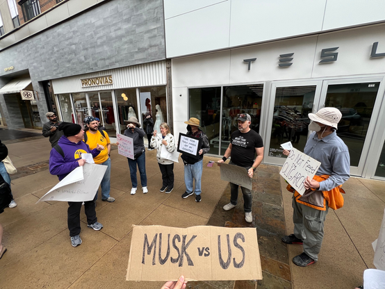 A variety of protestors outside a Tesla showroom, at the bottom of the camera is a cardboard sign which says "Musk vs US" 