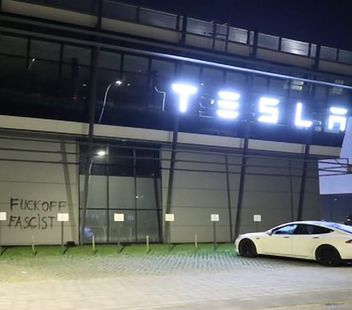 
A Tesla building at night, featuring a graffiti message reading "FUCK OFF FASCIST" on the side wall, with a parked white Tesla car nearby.