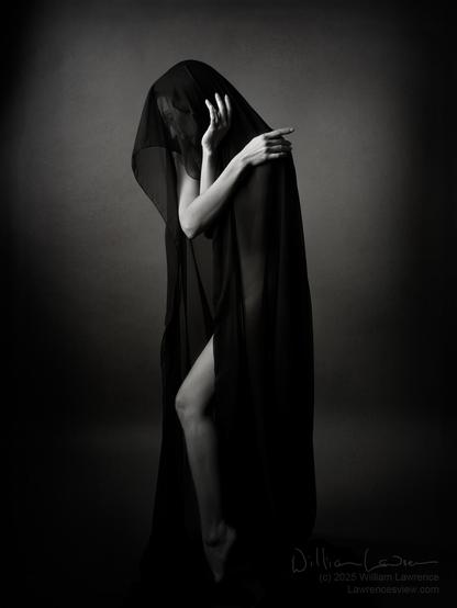 Black and white photograph, slightly toned, of model Zoe C West, posing nude but covered by a draping of sheer black fabric.  Only her arms, looking like arms of a clock, and one leg is clearly visible in this low key image.