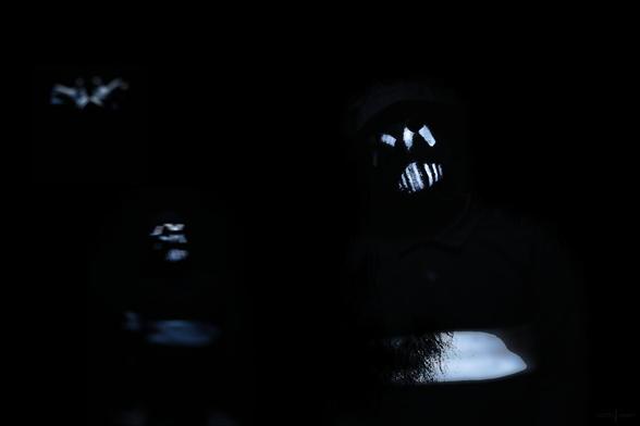 photo of the musical project "1 inch army". 1 inch army makes relectro music. the photo is a dark shot in front of a black background, wearing black clothings with some white details and stripes and white striped painted face masks.