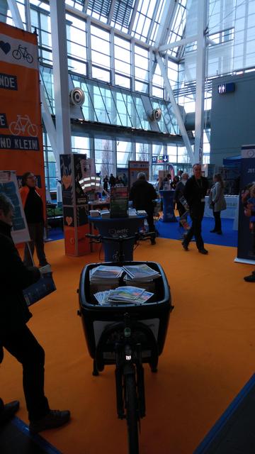 ADFC-Messestand auf der ABF. Auf orangenen und blauen Teppich laufen viele Messebesucher um die Prospektständer mit Radkarten aus ganz Norddeutschland r. Im Vordergrund ein Lastenrad mir sehr vielen Broschüren in der Kiste.