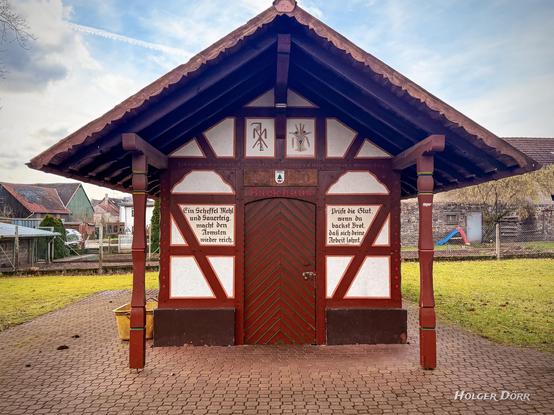 Das zweite Foto zeigt das Backhaus in Münster bei Laubach in der Frontansicht. Es ist ein charmantes kleines Fachwerkgebäude mit traditionellem Charakter. Die symmetrische Fassade wird von einer grünen Holztur mit rot-schwarzem Muster dominiert, die von zwei Schrifttafeln mit alten Sprüchen flankiert wird. Der linke Spruch betont den Wert von Mehl und Feuer für das Brotbacken, während der rechte zur Prüfung der Glut mahnt, um sicherzustellen, dass sich die Muhe lohnt. Über der Tür sind Backutensilien als Zierde angebracht, und die Inschrift „Backhaus" hebt die Funktion des Gebäudes hervor. Die Konstruktion mit ihrem weit überstehenden Dach wird von zwei hölzernen Stützen getragen, was dem Bauwerk ein uriges und einladendes Erscheinungsbild verleiht.
