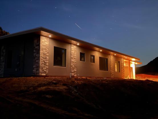House in progress with stucco and stone columns pictured from south-west at night, with accent lights in the front and brighter lights by the garage.