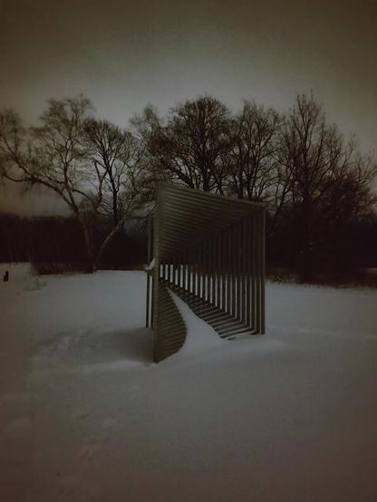 A dim snowy landscape with enough light bouncing off the ground to see through a slotted rectangular sculpture. There is a vignette effect, and bare branched trees frame the background. The sculpture is like a portal, where it gets smaller the deeper into the rectangle you look. A bit of snow rests on the slots that are parallel to the ground.