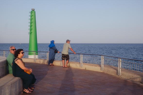 Photograph of the lighthouse in the port of Mataró. In the foreground there is a couple sitting, looking out to sea. In the background, next to the lighthouse, there is another couple standing, also looking out to sea, but in a different direction. My shadow is projected onto them (and I am also looking out to sea).

Fotografía del faro en el puerto de Mataró. En primer plano hay una pareja sentada, mirando al mar. Al fondo, junto al faro, hay otra pareja de pie, también mirando al mar, pero en otra dirección. Mi sombra se proyecta hasta ellos (y yo también estoy mirando el mar).