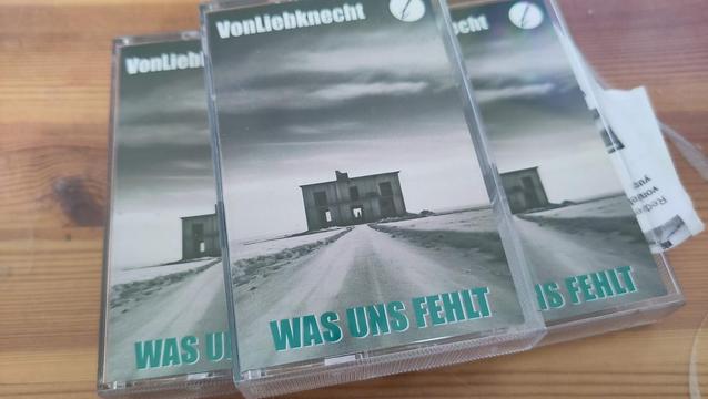 Das Foto zeigt drei Kassetten der vonLiebknecht EP "Was Uns Fehlt" auf einem Holztisch. Das Cover hat eine düstere, minimalistische Ästhetik: Ein verlassenes Haus steht in einer kargen, schneebedeckten Landschaft unter einem bewölkten Himmel. Der Bandname ist oben in weißer Schrift zu sehen, während der EP-Titel in grünen Großbuchstaben am unteren Rand steht. Die Kassetten sind in transparenten Plastikgehäusen verpackt, eine davon liegt leicht versetzt über den anderen. 