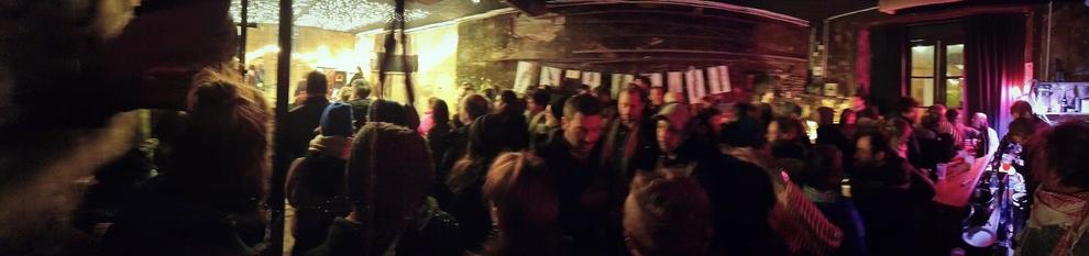 Photo panoramique d'un intérieur avec plein de monde. Les visages sont flous. à droite on peut comprendre qu'il y a un bar, et à gauche, plus de lumière avec une scène