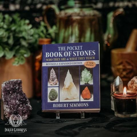 A copy of The Pocket Book of Stones is displayed on a stand next to an amethyst geode and votive candle. The cover has 5 photos of various crystals on the cover and is small enough to carry with you. Presented by Inked Goddess Creations.