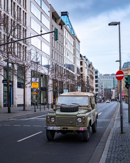 Ein alter, olivgrüner Geländewagen der Marke Land Rover steht auf einer mehrspurigen Straße in einer modernen Innenstadt. Der Wagen hat ein Reserverad auf der Motorhaube, ein weiches Stoffverdeck und ein deutsches Oldtimer-Kennzeichen. An der vorderen Stoßstange ist eine kleine britische Flagge angebracht. Die Straße ist von modernen Büro- und Geschäftshäusern mit großen Glasfassaden gesäumt. Die Bäume entlang der Straße sind kahl, was auf eine winterliche Jahreszeit hindeutet. Rechts im Bild befindet sich ein „Einfahrt verboten“-Verkehrsschild. Weiter hinten sind einige Fußgänger und Fahrzeuge zu sehen, darunter ein Taxi. 