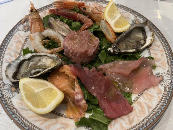 "Gran Crudo" : Un magnifique plateau de fruits de mer cru sarde avec huîtres, crevettes et carpaccio de poisson, accompagné de quartiers de citron. A beautiful seafood platter featuring oysters, shrimp, and fish carpaccio, served with lemon wedges.