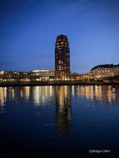 Ein nächtliches Stadtbild von Frankfurt am Main. Im Zentrum des Fotos steht das markante „Main Plaza“ mit seiner beleuchteten Fassade, das sich in den ruhigen Wassern des Mains spiegelt. Der Himmel zeigt ein tiefes Blau der Dämmerung, während Straßenlaternen und umliegende Gebäude ein warmes Licht auf das Ufer werfen. Die Szenerie wirkt ruhig und stimmungsvoll, eine alternative Perspektive auf die Skyline von Frankfurt.