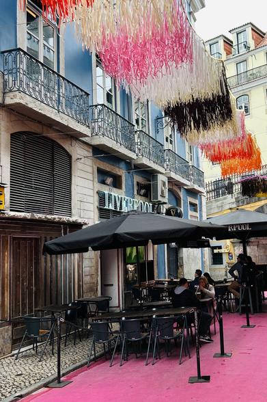 A narrow street features a colorful overhead decoration, with pink, orange, and brown elements. Below, there are outdoor tables and chairs beneath a black umbrella, with patrons seated.