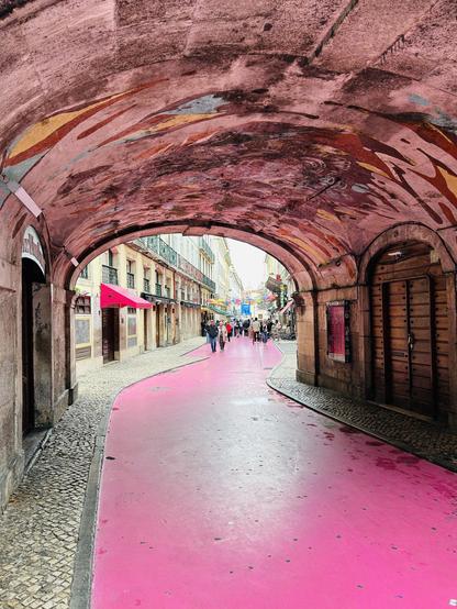 A vibrant pink pathway runs through an arched walkway, lined with buildings displaying colorful facades. People are walking along the street, and decorative murals adorn the arch above, creating a lively atmosphere.