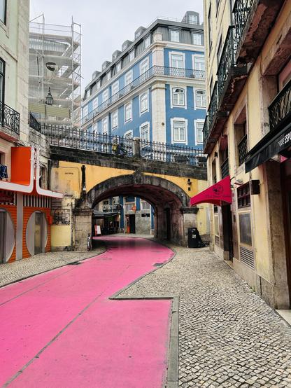 A narrow, winding street features a vibrant pink pathway bordered by cobblestones, with historic buildings on either side. An archway leads to a different area, and construction scaffolding is visible on one building.