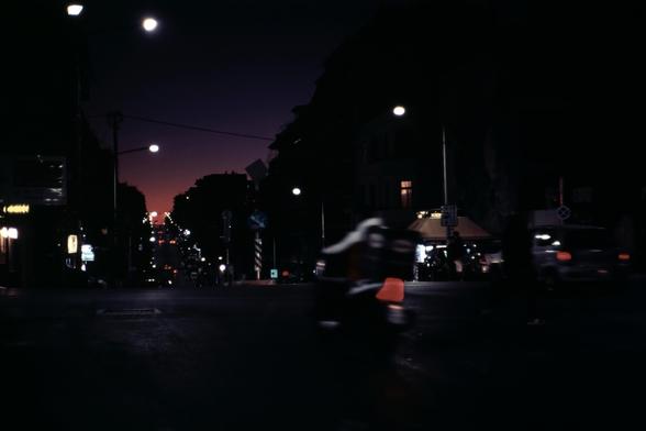A motor scooter rides down a darkly lit road in Athens, Greece towards a deep pink and purple sunset. It is the last light of day. 