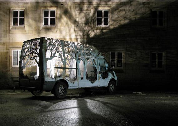 Photo d'un camion blanc de nuit, garé devant la façade d'une maison. Toute sa carrosserie est découpée de sorte à représenter la silhouette d'une forêt et en son centre on peut y voir une lumière blanche qui en sort, le camion ressemble à une grande lanterne sur roues.