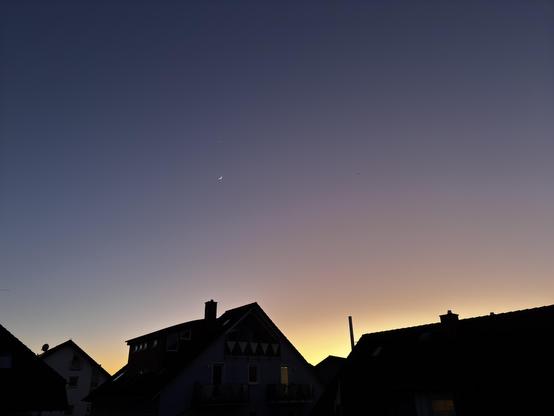 Motiv:
Das Bild zeigt eine Stadtsilhouette mit mehreren Wohnhäusern im unteren Bereich. Der Himmel nimmt den größten Teil des Bildes ein und zeigt einen Farbverlauf von dunklem Blau zu warmem Gelb. Eine schmale Mondsichel und ein heller Himmelskörper sind sichtbar.

Stimmung & Wirkung:
Die Aufnahme wirkt ruhig und weitläufig. Die Kontraste zwischen den dunklen Gebäuden und dem Himmel betonen die Silhouetten. Die Himmelsfarben verleihen dem Bild eine natürliche Tiefe.

Technische Details:
Bildformat: Landschafts- bzw. Himmelsfotografie.

Farbe: Natürlicher Farbverlauf von Dunkelblau zu Gelb.

Schärfentiefe: Tiefe Schärfentiefe, klare Darstellung der Gebäude und Himmelskörper.

Lichtverhältnisse: Natürliches Dämmerlicht mit sanften Übergängen.