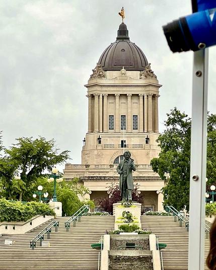 A back view photo of the Manitoba Legislative Building,taken from a boat on the Assiniboine River. The Golden Boy sits atop the building. At the bottom of the many stairs leading to the River bank is a bronze statue of Louis Riel. Trees and lamp posts line the sides of the stairs. 