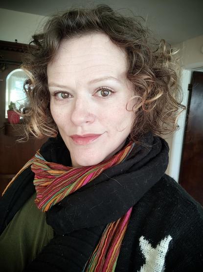 Photo of me, middle-aged white woman with brown eyes and dark blond, chin-length curly hair. The curls are especially soft and bouncy today. I'm wearing two thin scarves, one black and one multi-colored striped. Top is dark olive green with a knit cardigan over it. Cardigan is black with large  white stars.